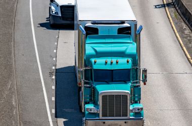 Camion industriale acquamarina con motrice a cabina estesa per il riposo del conducente, che trasporta merci in un rimorchio frigorifero, in corsa su una strada autostradale davanti a un altro camion articolato.